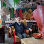 Gaetano Pesce seduto nel suo studio circondato da sculture e oggetti in resina dai colori accesi, tra cui un grande vaso rosso con un volto