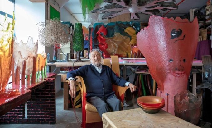 Gaetano Pesce seduto nel suo studio circondato da sculture e oggetti in resina dai colori accesi, tra cui un grande vaso rosso con un volto