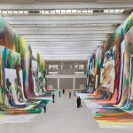 Grandi teli dipinti con campiture fluide multicolori sospesi lungo le pareti di una sala espositiva, con visitatori nel corridoio centrale