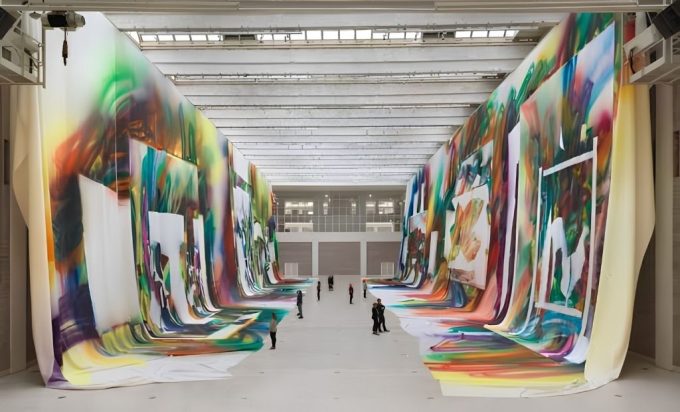 Grandi teli dipinti con campiture fluide multicolori sospesi lungo le pareti di una sala espositiva, con visitatori nel corridoio centrale