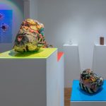 Veduta d'allestimento con due sculture ceramiche bulbose dai motivi policromi su piani in acrilico giallo e azzurro, e una stampa blu iridescente alla parete
