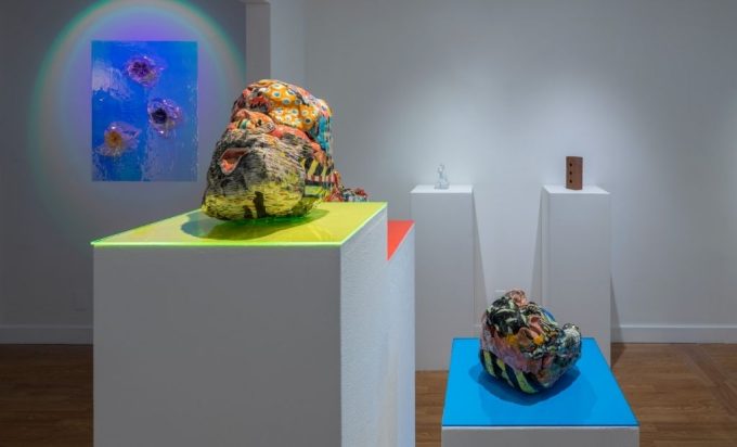 Veduta d'allestimento con due sculture ceramiche bulbose dai motivi policromi su piani in acrilico giallo e azzurro, e una stampa blu iridescente alla parete