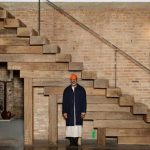 Theaster Gates in piedi davanti a una scala monumentale costruita con travi di legno di recupero, in uno spazio industriale in mattoni