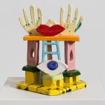Scultura in ceramica colorata con mani, occhio e labbra rosse su una struttura geometrica